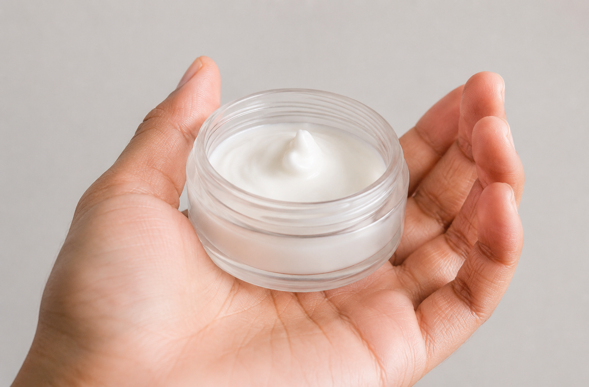A hand holding a small clear jar filled with white cream against a plain light gray background.
