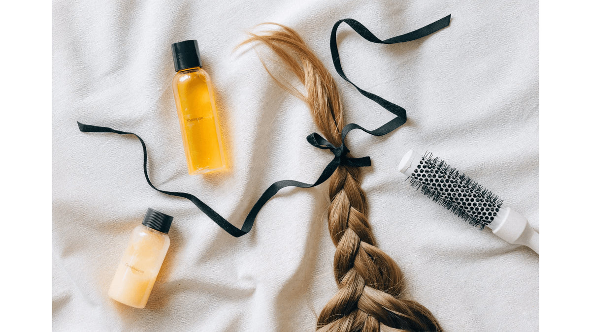 Two small bottles of hair care liquid with black caps, a long blonde braid tied with a black ribbon, and a white round hairbrush arranged on a white fabric background.